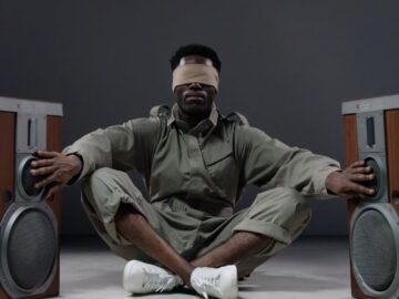 Blindfolded man seated with arms outstretched touching speakers on each side, exploring sound.