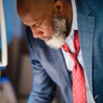Close-up of a distinguished bearded man in a suit leaning over, focused and thoughtful.
