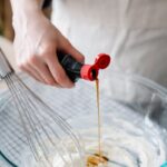 Close-up of a hand pouring vanilla extract into a cream mixture in a glass bowl with a whisk.