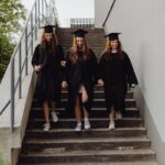 Three young women in graduation gowns descend outdoor steps, celebrating their academic achievements.