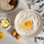 Flat lay photo of baking ingredients including flour, butter, and eggs on a white surface.