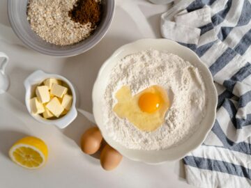 Flat lay photo of baking ingredients including flour, butter, and eggs on a white surface.