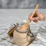 A close-up shot of a hand scooping oats from a glass jar on a striped cloth in a kitchen setting.