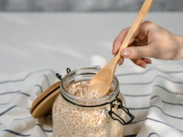 A close-up shot of a hand scooping oats from a glass jar on a striped cloth in a kitchen setting.