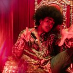 Man in vibrant outfit and afro hairstyle posing in a creative indoor setting.