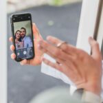 Close-up of hands holding a smartphone showing a couple taking a selfie outdoors.