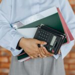 Close-up of a person holding a calculator and folders against a brick wall background.