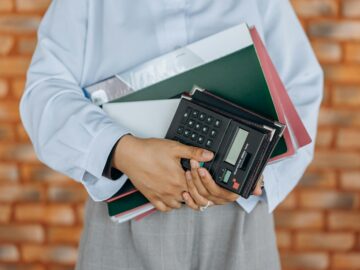 Close-up of a person holding a calculator and folders against a brick wall background.
