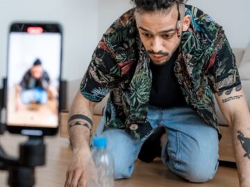 A man filming a bottle flip challenge on a smartphone indoors, capturing a trending social media moment.