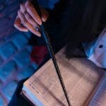 Close-up of a person holding a wand while reading an open book with focused lighting.