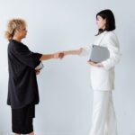 Two businesswomen shaking hands in an office setting, symbolizing partnership and teamwork.
