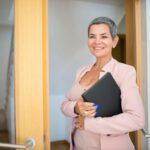 Smiling female real estate agent holding a black folder inside a modern building.