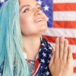 A woman with blue hair wrapped in an American flag, gazing upwards with hands in prayer position.