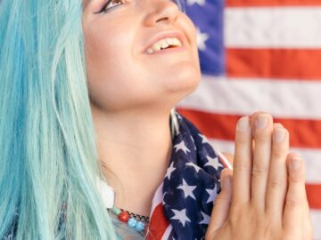 A woman with blue hair wrapped in an American flag, gazing upwards with hands in prayer position.