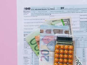 A vibrant flat lay of a calculator, euro banknotes, and US tax forms on a pink background.