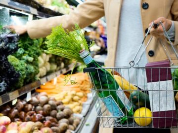Woman selecting fresh produce in a supermarket with a basket full of groceries.