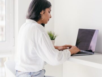 Woman sitting on stool writing on laptop, showcasing home study setup.
