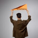 Back view of a person holding a kindness sign on a gray background, symbolizing compassion and empathy.
