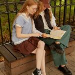 Two women sitting on a wooden bench in the park, enjoying a book together on a sunny day.