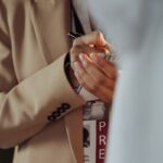 Journalist in a beige coat taking notes with a visible press badge.