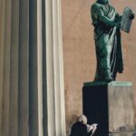Woman reading in sunlight next to a grand stone statue and column.