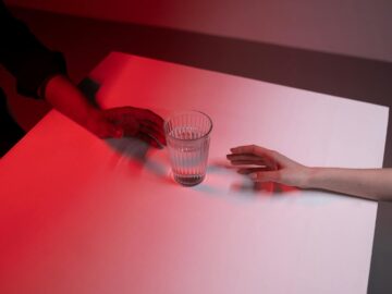 Two diverse hands reaching towards a glass of water on a table with dramatic red lighting.