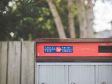 A Canada Post mailbox with colorful label outdoors with trees in the background.