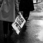 grayscale photo of person holding white and blue no smoking sign