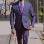 man in blue suit walking on the sidewalk near metal fence and parked vehicle