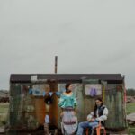 Portrait of a couple in traditional attire outside a rustic dwelling under a gray sky.