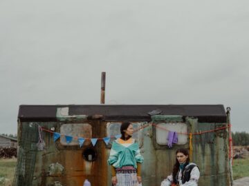 Portrait of a couple in traditional attire outside a rustic dwelling under a gray sky.