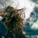 A woman with disheveled hair caught in strong wind against a stormy backdrop, capturing motion and chaos.