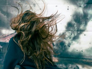 A woman with disheveled hair caught in strong wind against a stormy backdrop, capturing motion and chaos.