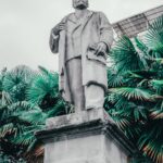 Low angle view of Lenin statue surrounded by lush tropical plants, under an overcast sky.
