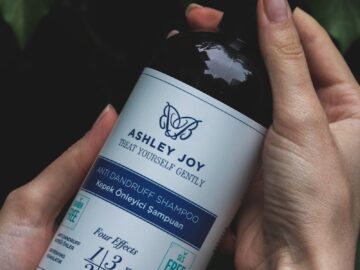 Close-up of hands holding an Ashley Joy anti-dandruff shampoo bottle with nature background.