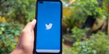 Hand holding a smartphone with the Twitter app open, outdoors with greenery in the background.