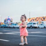 Smiling toddler in a pink outfit standing in an urban parking lot with cars and neon lights.