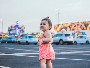 Smiling toddler in a pink outfit standing in an urban parking lot with cars and neon lights.