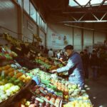 A bustling indoor market with vibrant fresh fruits and vegetables on display and people shopping.