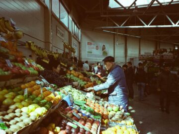 A bustling indoor market with vibrant fresh fruits and vegetables on display and people shopping.