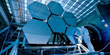 Engineers in protective suits work on telescopic mirrors in a high-tech lab.