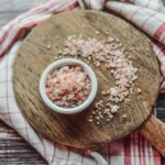 A bowl of pink Himalayan salt on a wooden board surrounded by a checkered cloth, viewed from above.