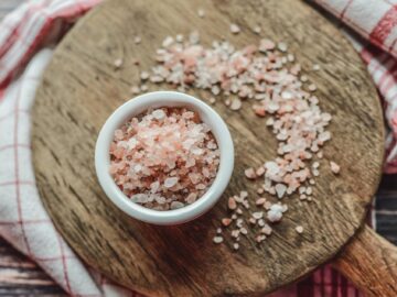 A bowl of pink Himalayan salt on a wooden board surrounded by a checkered cloth, viewed from above.