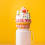 Vibrant milkshake adorned with a donut, whipped cream, and candy against a yellow background.