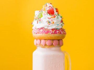 Vibrant milkshake adorned with a donut, whipped cream, and candy against a yellow background.