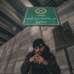 A man poses under a Persian language no parking sign in an urban setting.