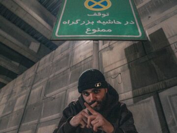A man poses under a Persian language no parking sign in an urban setting.
