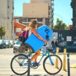 A woman on a bicycle carries a surfboard and backpack through a bustling city street.