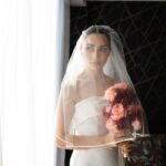 A beautiful bride in a strapless gown holding a bouquet by a window, exuding elegance.