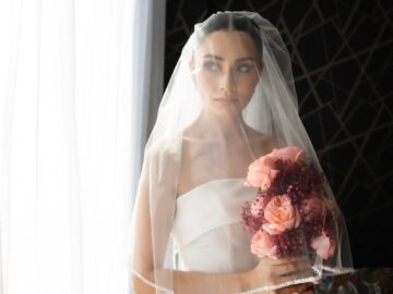 A beautiful bride in a strapless gown holding a bouquet by a window, exuding elegance.